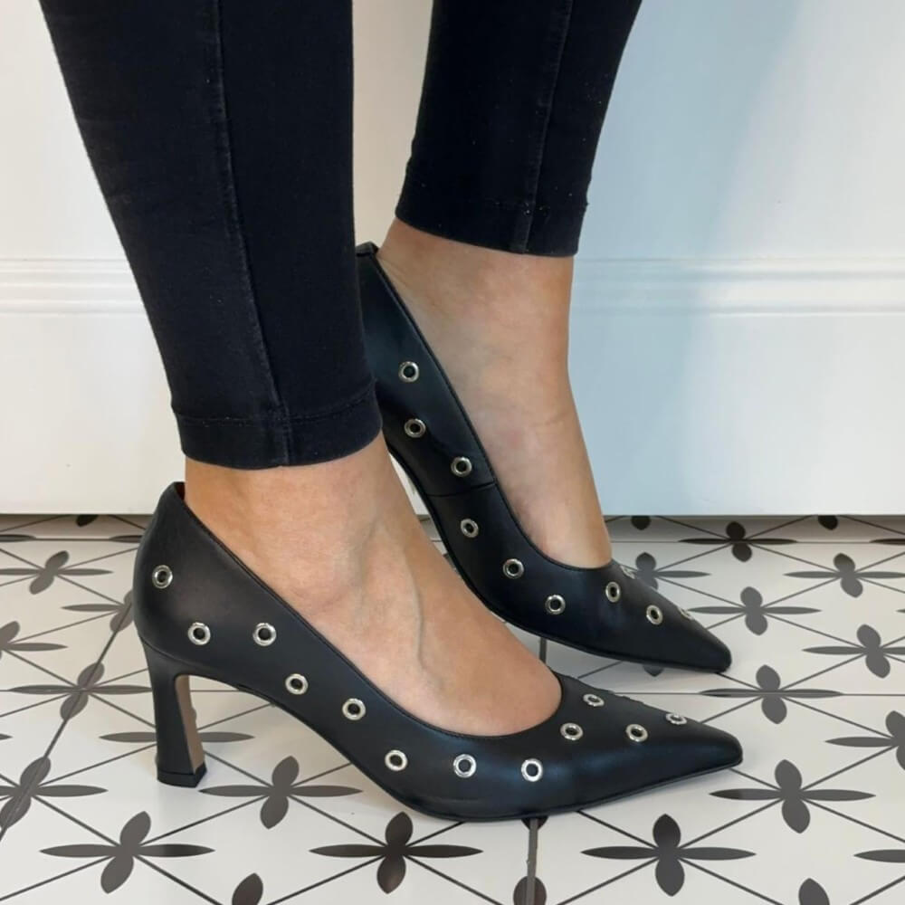 Black high-heeled shoes with decorative studs worn by a person on a patterned floor. Ángel Alarcón Adriene 25557 Leather Court Shoe-BLACK