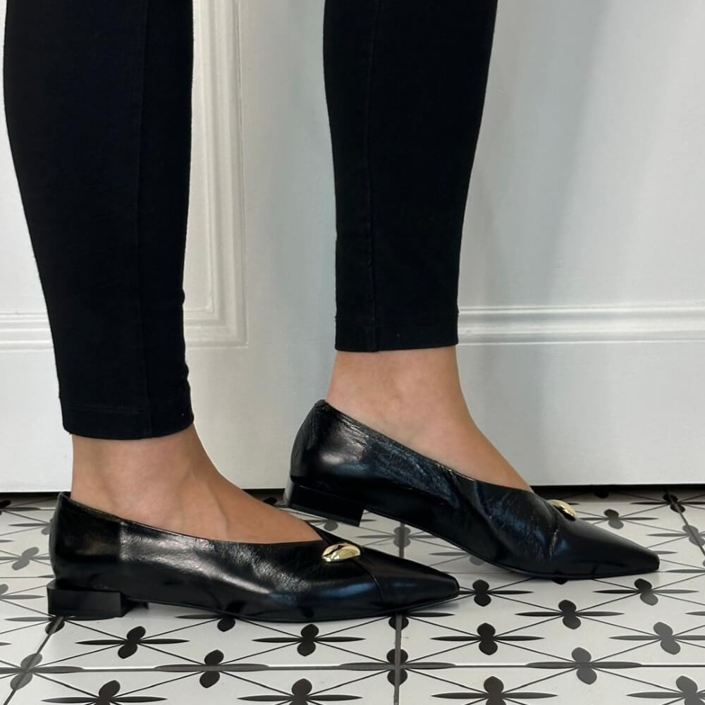 A person wearing Angel Alarcon black pointed-toe flats with small gold-tone decorative accents, shown from the calves down against a white wall and black-and-white patterned tile floor.