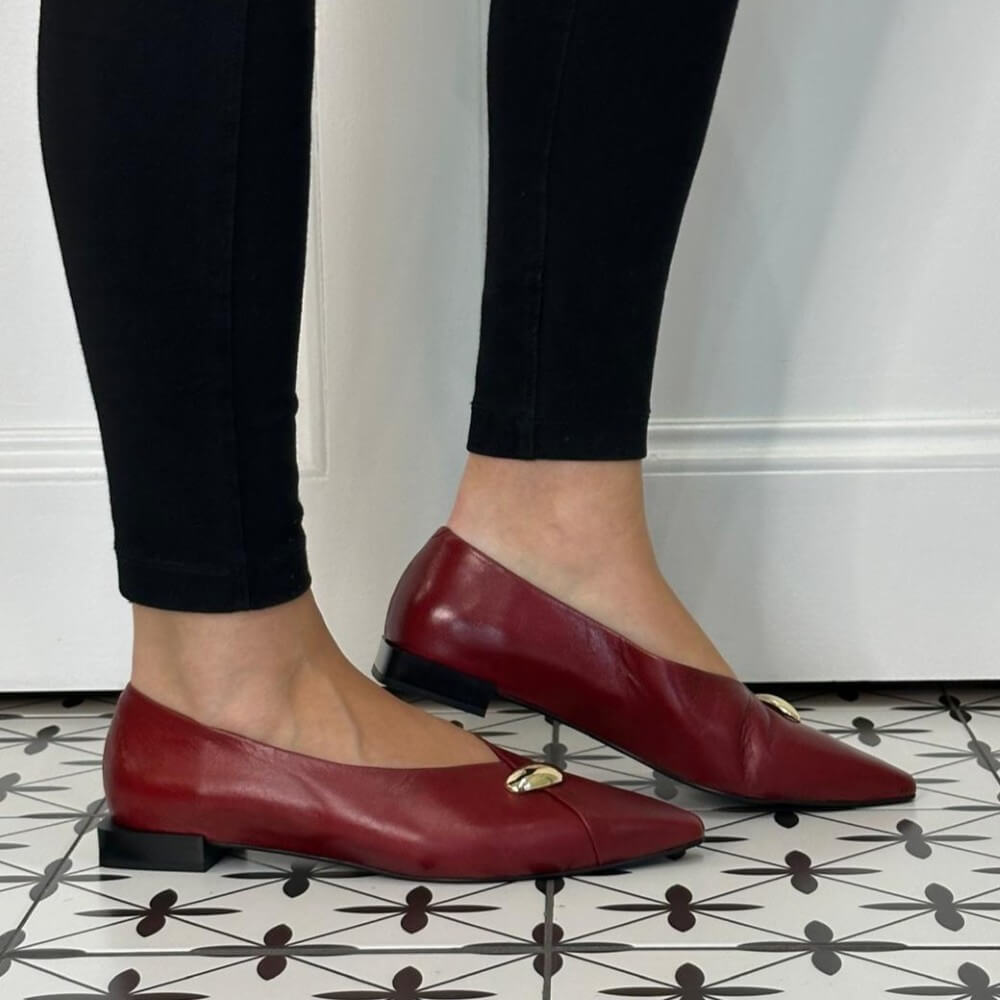 A person wearing black leggings and Angel Alarcon red leather pointed-toe flats with small gold-tone decorative accents, standing on a black-and-white patterned tile floor against a white wall.
