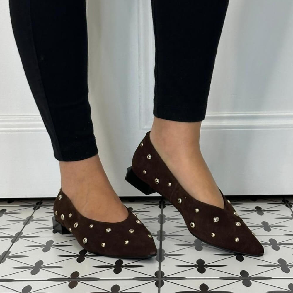 A person wearing Angel Alarcon dark brown suede pointed-toe flats with silver stud embellishments, paired with black leggings, standing on a black and white geometric patterned tile floor.