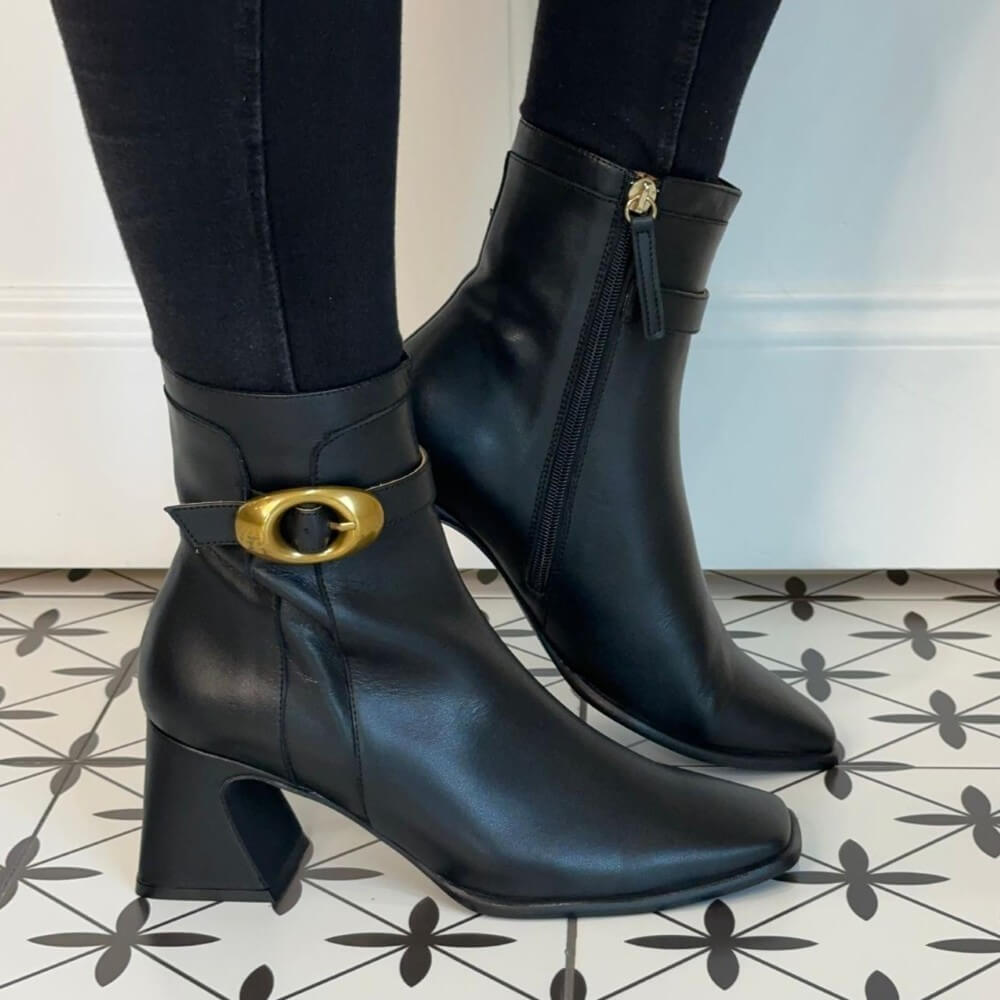 Black ankle boots with gold buckle worn indoors on a patterned floor. Ángel Alarcón Sandrie 25570 Ankle Boot-BLACK
