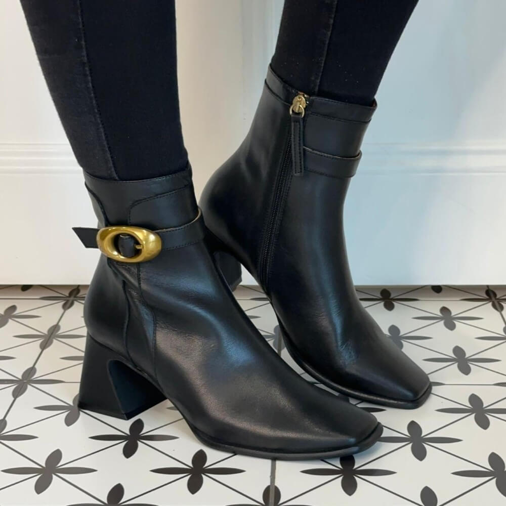 Black ankle boots with gold buckle worn indoors on a patterned floor. Ángel Alarcón Sandrie 25570 Ankle Boot-BLACK