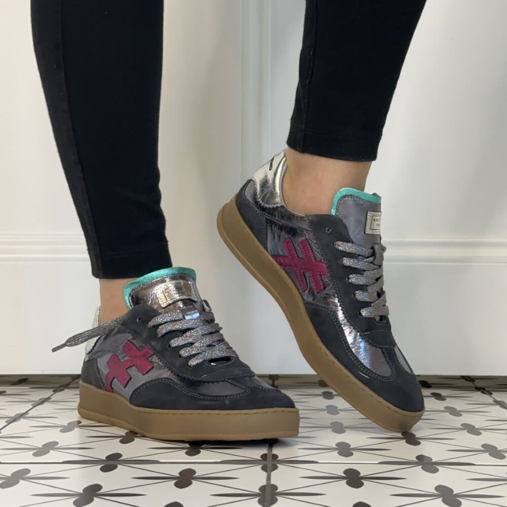 A person wearing black leggings and Another Trend grey low-top sneakers with metallic silver overlays, a pink cross logo, and gum soles, standing on a patterned tile floor against a white wall.