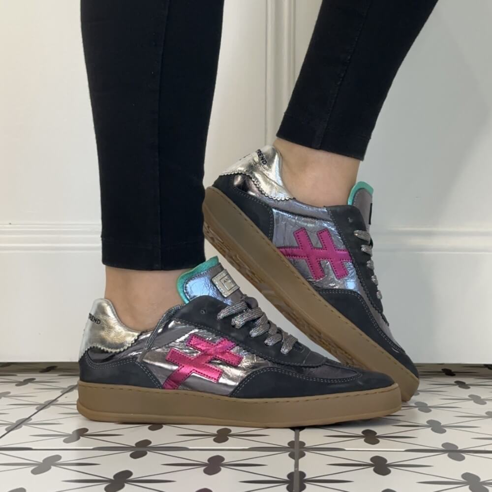 A person wearing black leggings and Another Trend grey low-top sneakers with metallic silver overlays, a pink cross logo, and gum soles, standing on a patterned tile floor against a white wall.