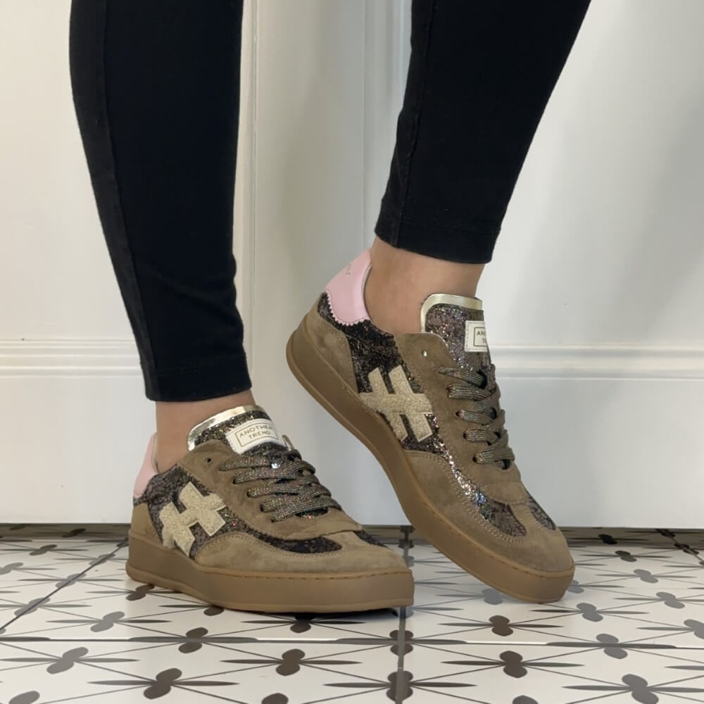 A person wearing black leggings and Another Trend brown low-top sneakers with pink heel tabs and glittery beige cross logos, standing on a patterned tile floor against a white wall.
