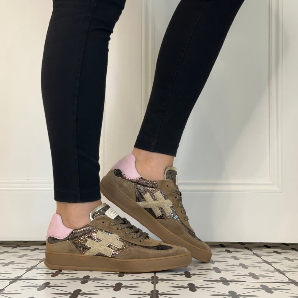 A person wearing black leggings and Another Trend brown low-top sneakers with pink heel tabs and glittery beige cross logos, standing on a patterned tile floor against a white wall.
