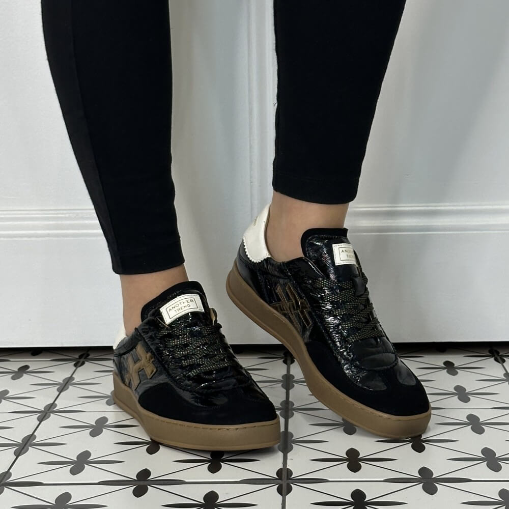 A person wearing Another Trend black patent leather sneakers with gold metallic '#' logos, white heel tabs, and tan gum soles, paired with black leggings on a patterned tile floor.