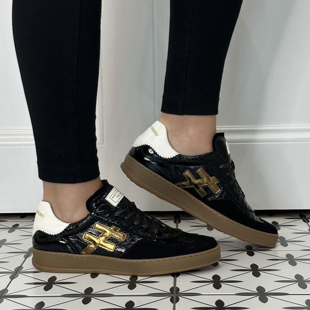 A person wearing Another Trend black patent leather sneakers with gold metallic '#' logos, white heel tabs, and tan gum soles, paired with black leggings on a patterned tile floor.