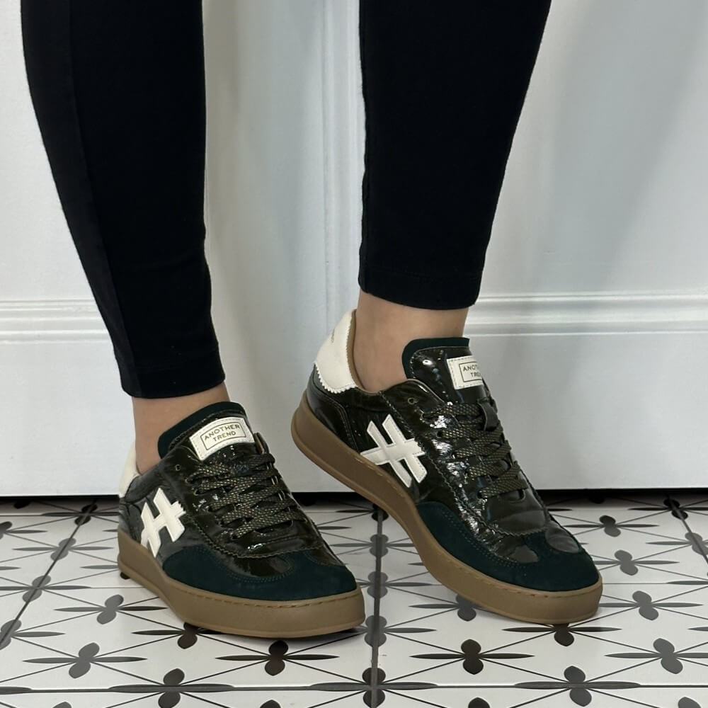 A person wearing black leggings and Another Trend dark green patent leather sneakers with white '#' side stripes, cream heel tabs, and brown gum soles, standing on a black-and-white patterned tile floor against a white panelled wall.
