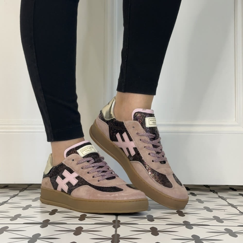 A pair of Another Trend low-top sneakers worn with black leggings, shown from the side on a patterned tile floor against a white wall. The shoes feature a dusty pink suede base, black glitter panels, a light pink cross logo, and metallic gold heel accents.