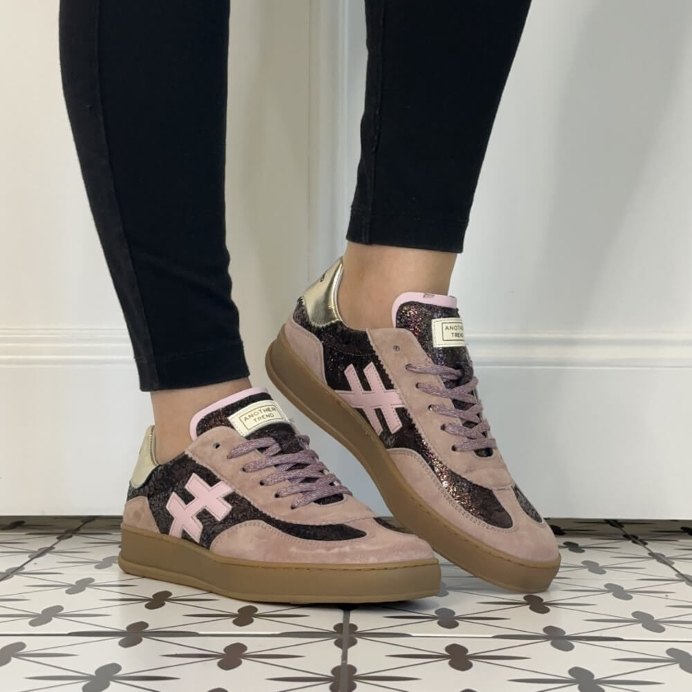 A pair of Another Trend low-top sneakers worn with black leggings, shown from the side on a patterned tile floor against a white wall. The shoes feature a dusty pink suede base, black glitter panels, a light pink cross logo, and metallic gold heel accents.
