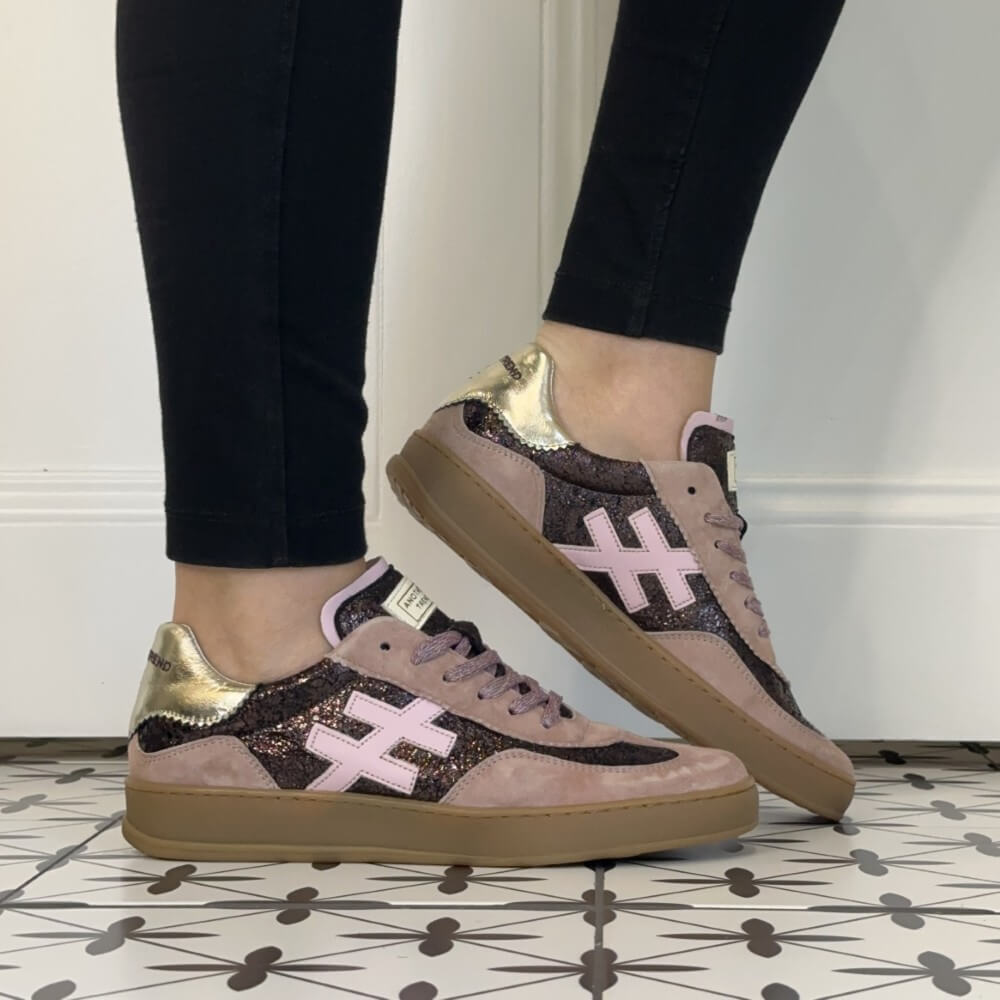 A pair of Another Trend low-top sneakers worn with black leggings, shown from the side on a patterned tile floor against a white wall. The shoes feature a dusty pink suede base, black glitter panels, a light pink cross logo, and metallic gold heel accents.