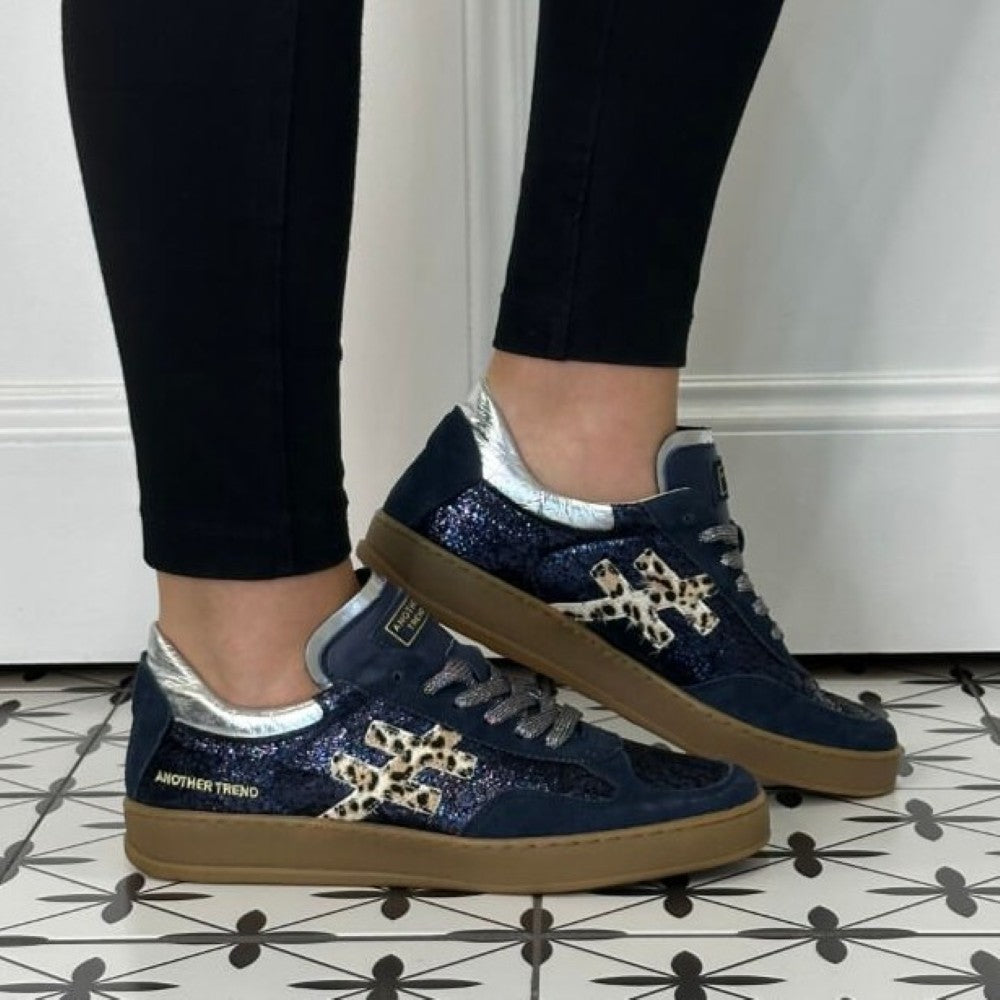A person wearing Another Trend navy blue glitter sneakers with leopard print overlays and silver accents, paired with black leggings, standing on a black-and-white patterned tile floor.