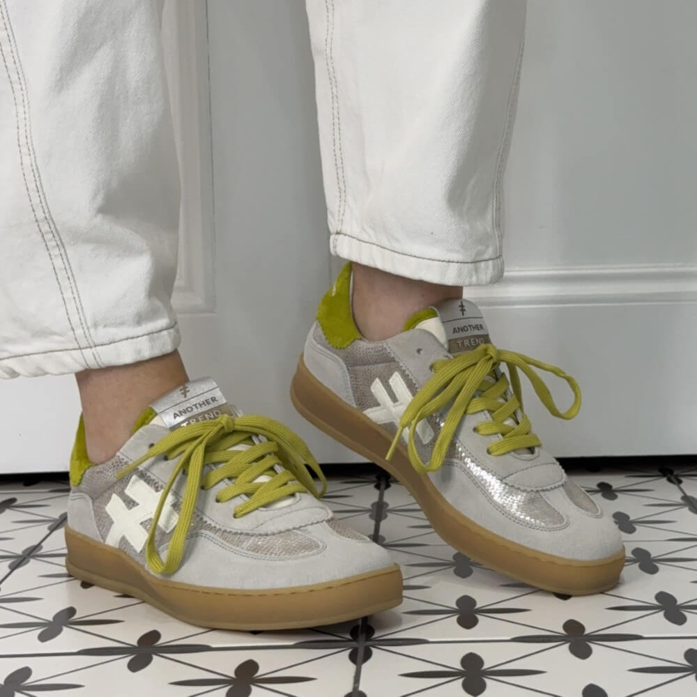 Champagne sneakers with yellow laces worn by a person on a patterned floor.