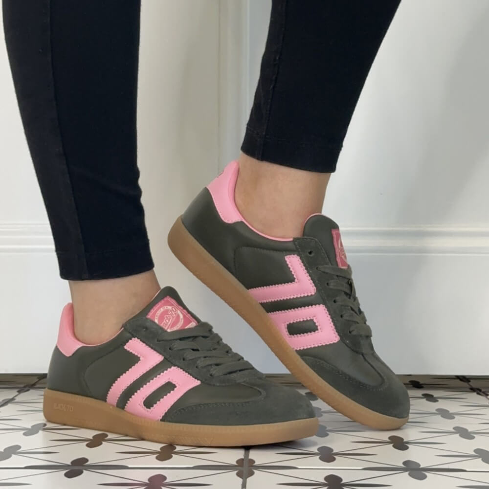 A person wearing dark green and pink Back 70 sneakers with black leggings, standing on a patterned tile floor against a white wall.