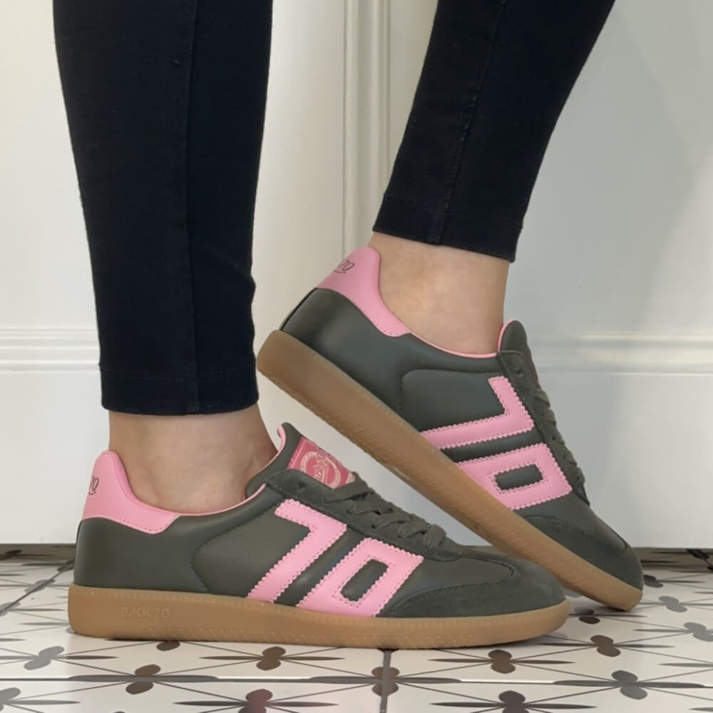 A person wearing dark green and pink Back 70 sneakers with black leggings, standing on a patterned tile floor against a white wall.