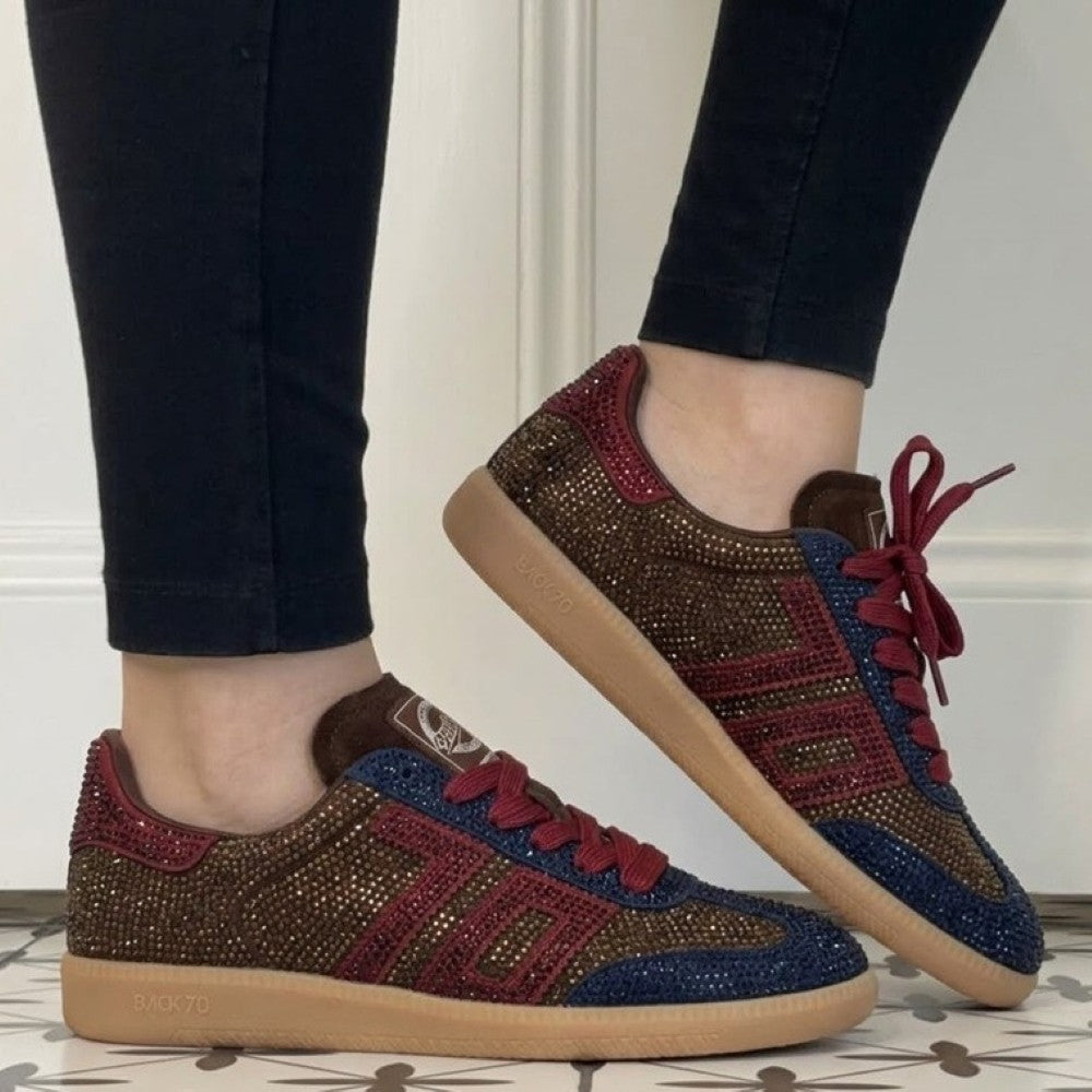A pair of Back 70 low-top sneakers being worn, shown from the side and back. The shoes feature a brown suede base heavily embellished with gold rhinestones, with dark blue rhinestone panels on the sides and red rhinestone accents on the toe and laces.