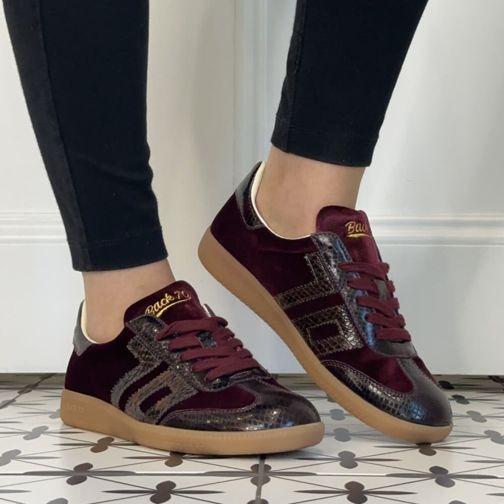 A person wearing Back 70 dark burgundy velvet and snakeskin-patterned sneakers with tan gum soles, paired with black pants, standing on a patterned tile floor.