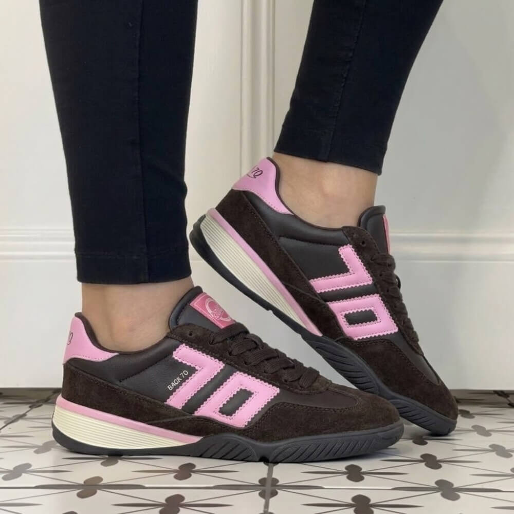 A person wearing Back 70 dark brown suede and leather sneakers with bright pink accents and cream midsoles, paired with black leggings, standing on a patterned tile floor
