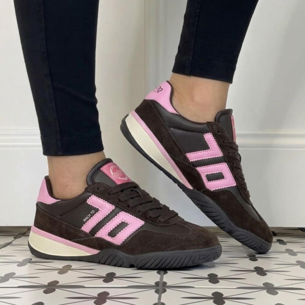 A person wearing Back 70 dark brown suede and leather sneakers with bright pink accents and cream midsoles, paired with black leggings, standing on a patterned tile floor