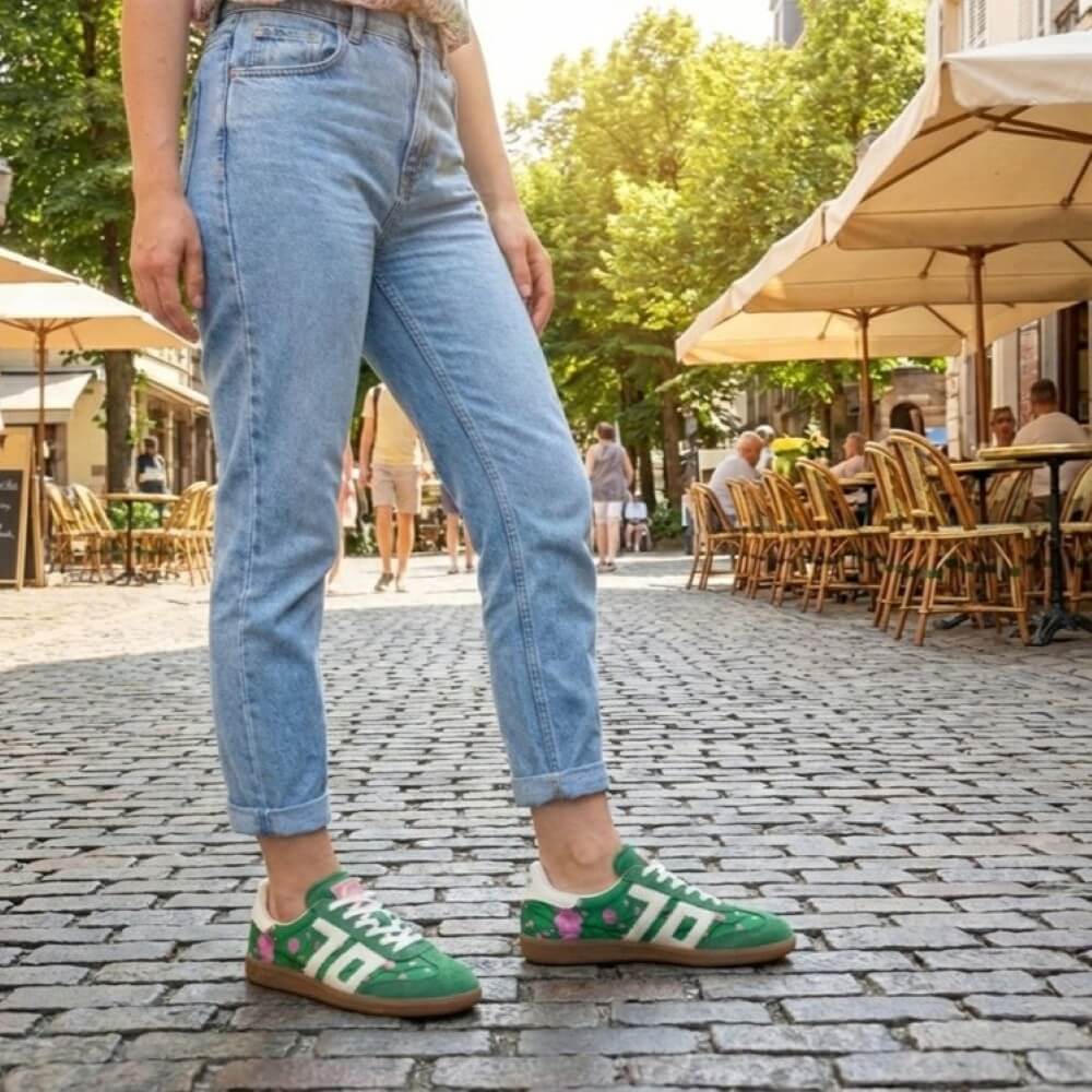 Person wearing Back 70 green sneakers and blue jeans standing on a cobbled street with outdoor seating in the background.