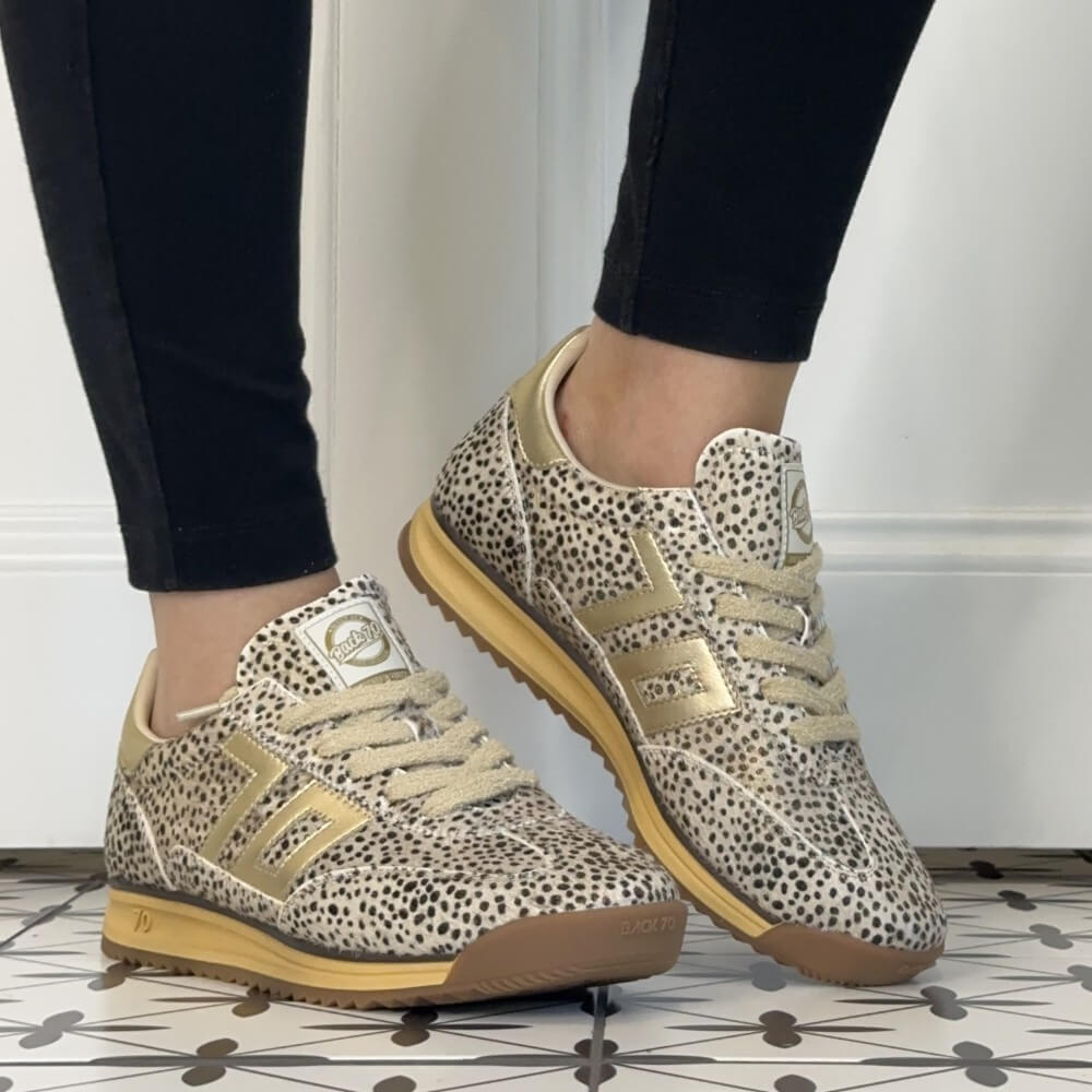 A pair of Back 70 beige and black leopard-print sneakers with metallic gold accents, worn with black leggings on a patterned tile floor.