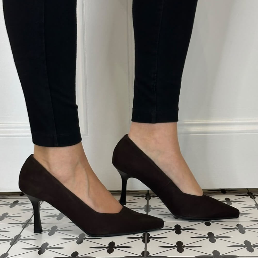 A pair of Bioeco dark brown pointed-toe high heels worn with black skinny jeans, shown on a patterned black and white tile floor against a white wall.