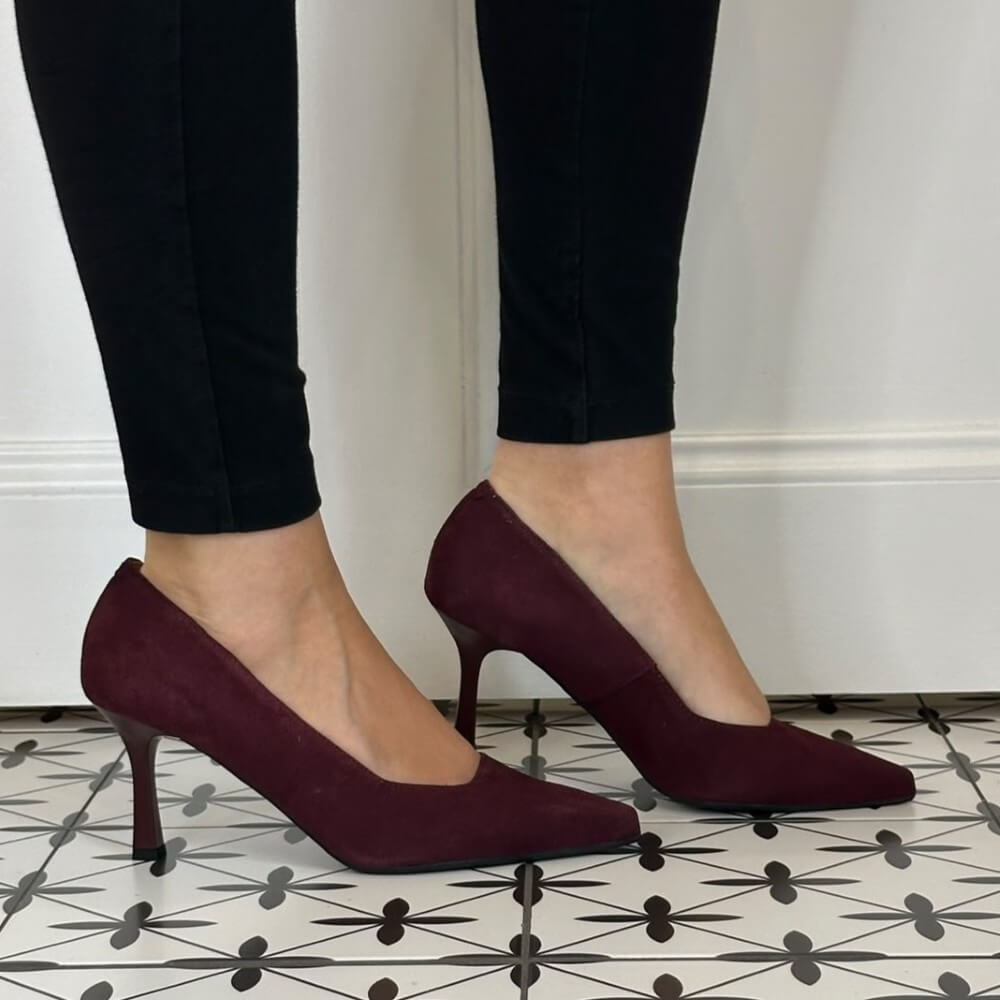 A person wearing Bioeco pointed-toe heeled pumps in deep burgundy suede, paired with black pants, standing on a black and white patterned tile floor.