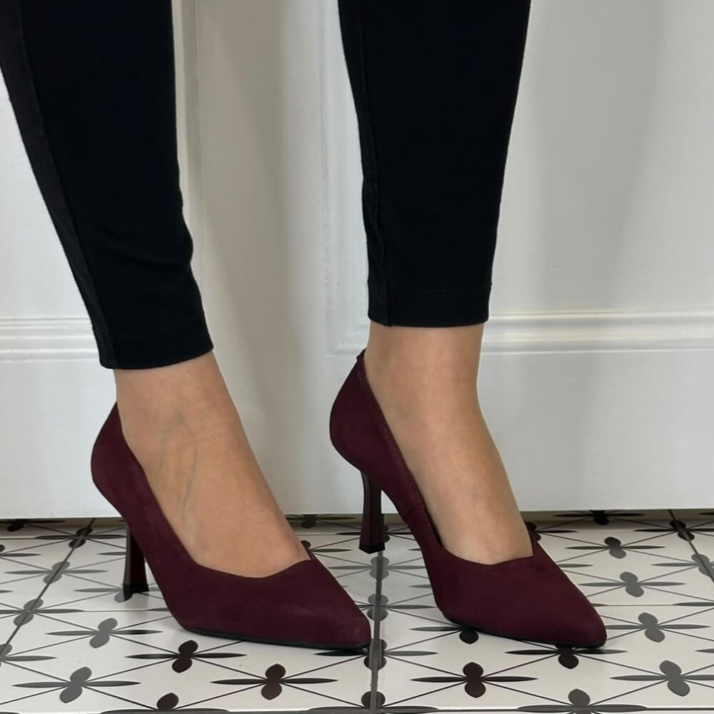 A person wearing Bioeco pointed-toe heeled pumps in deep burgundy suede, paired with black pants, standing on a black and white patterned tile floor.