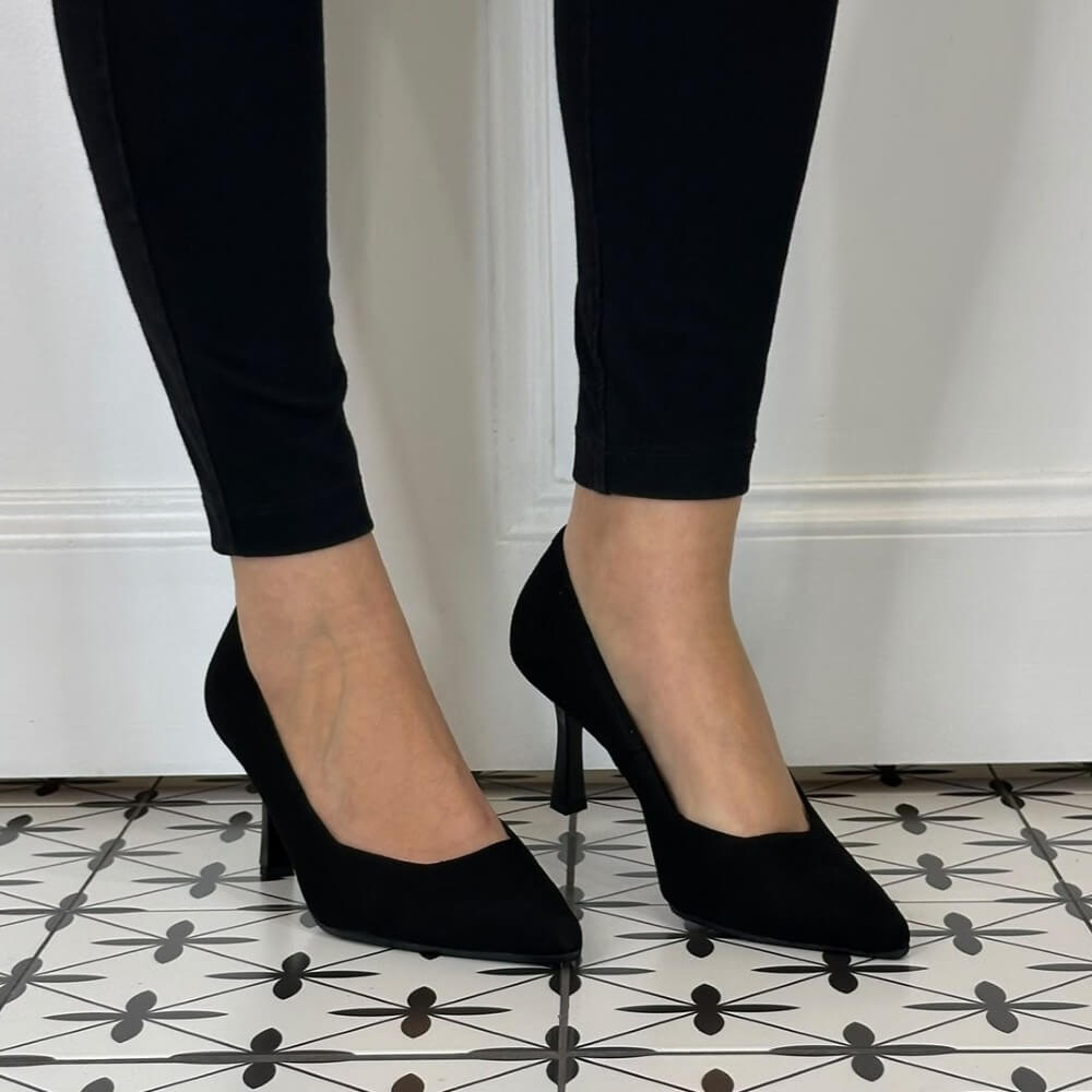 Bioeco Black pointed-toe high heels with slim stiletto heels, shown on a model wearing black pants against a white panelled wall and patterned tile floor.