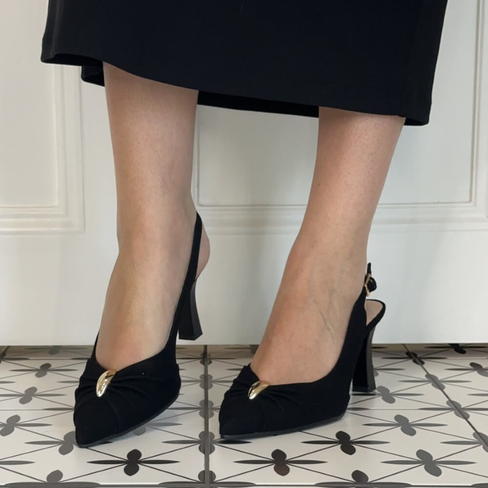A person wearing Bioeco black slingback pointed-toe pumps with gold-tone buckle details, shown from the knees down against a white paneled wall and patterned tile floor.