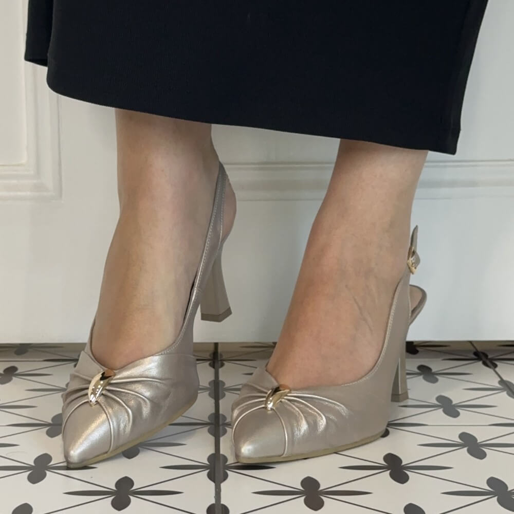 A person wearing Bioeco Gold slingback pointed-toe pumps with gold-tone buckle details, shown from the knees down against a white panelled wall and patterned tile floor.