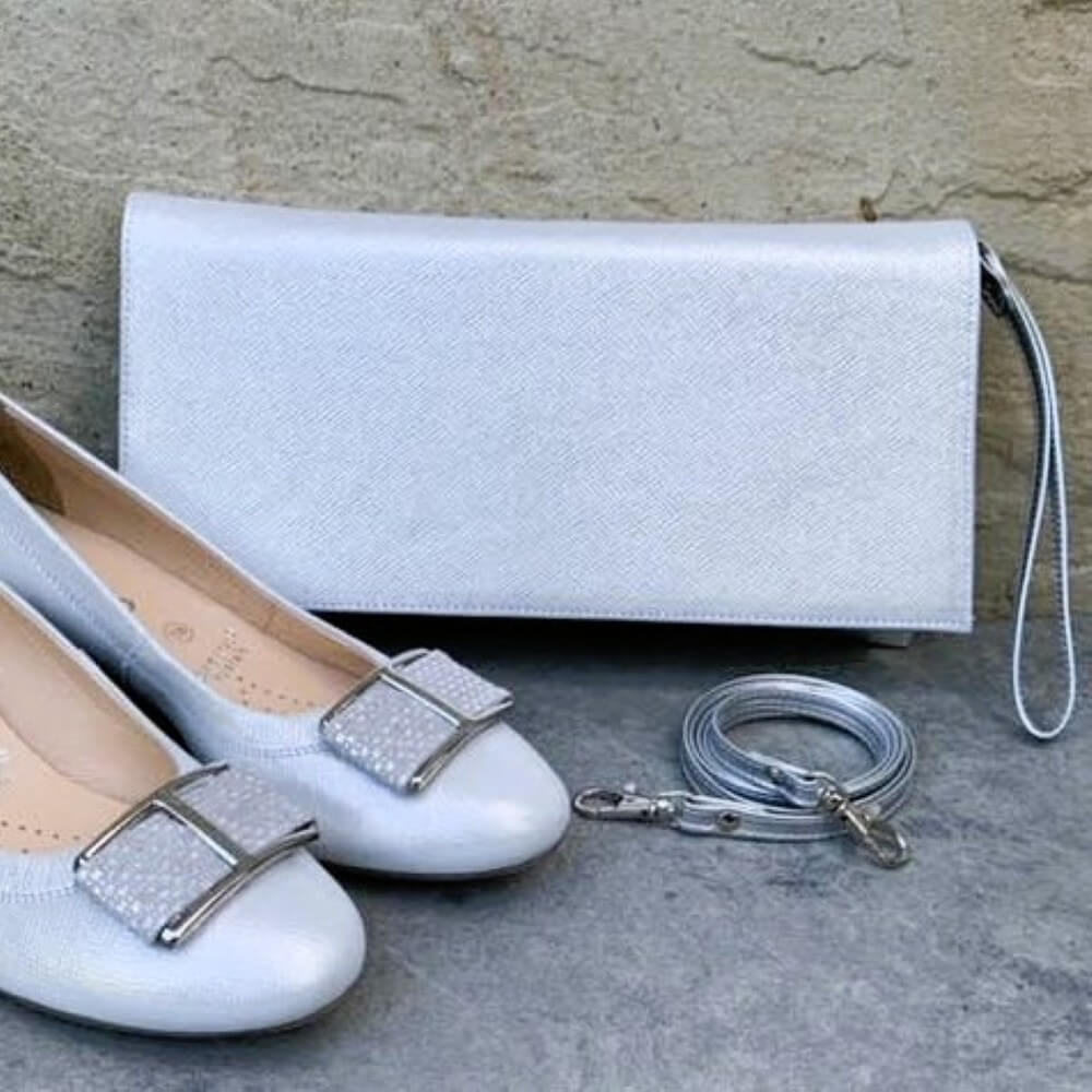 Pair of silver shoes with silver buckles and a matching clutch bag on a stone surface.