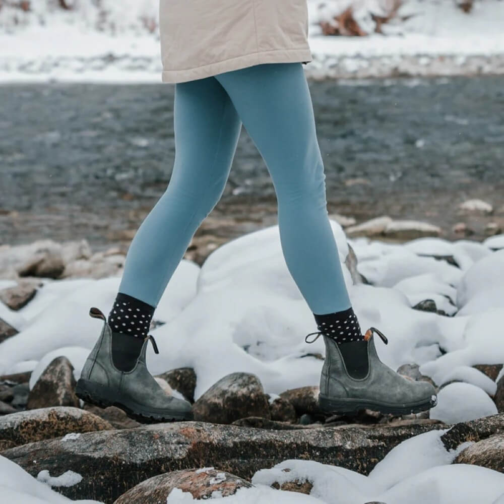 Person wearing blue leggings and black boots standing on a snowy landscape with water in the background.