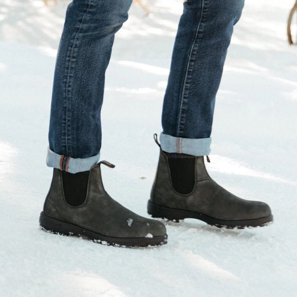 Person wearing black Chelsea boots and blue jeans standing on a snowy ground.