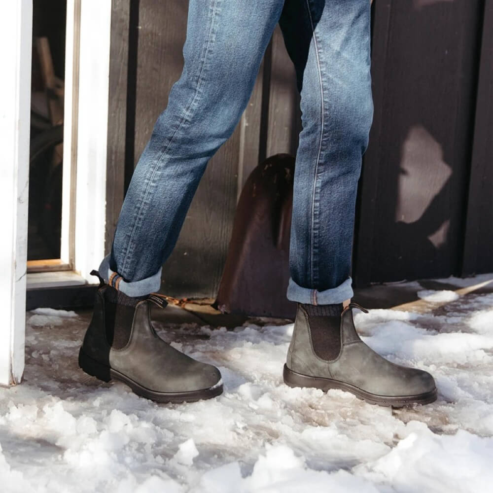 Person wearing lack Chelsea boots and blue jeans standing on a snowy ground.