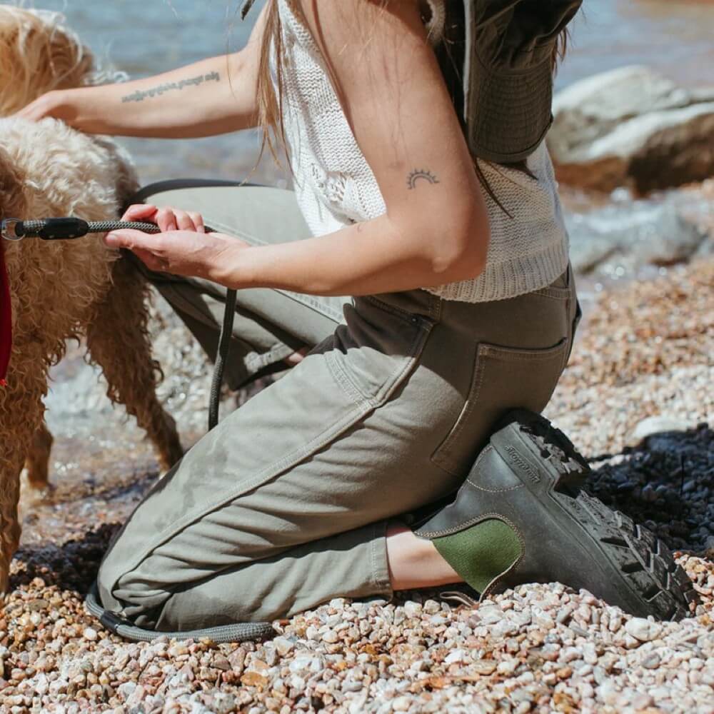 Person kneeling on a rocky beach with a dog, wearing brown green boots and khaki pants.