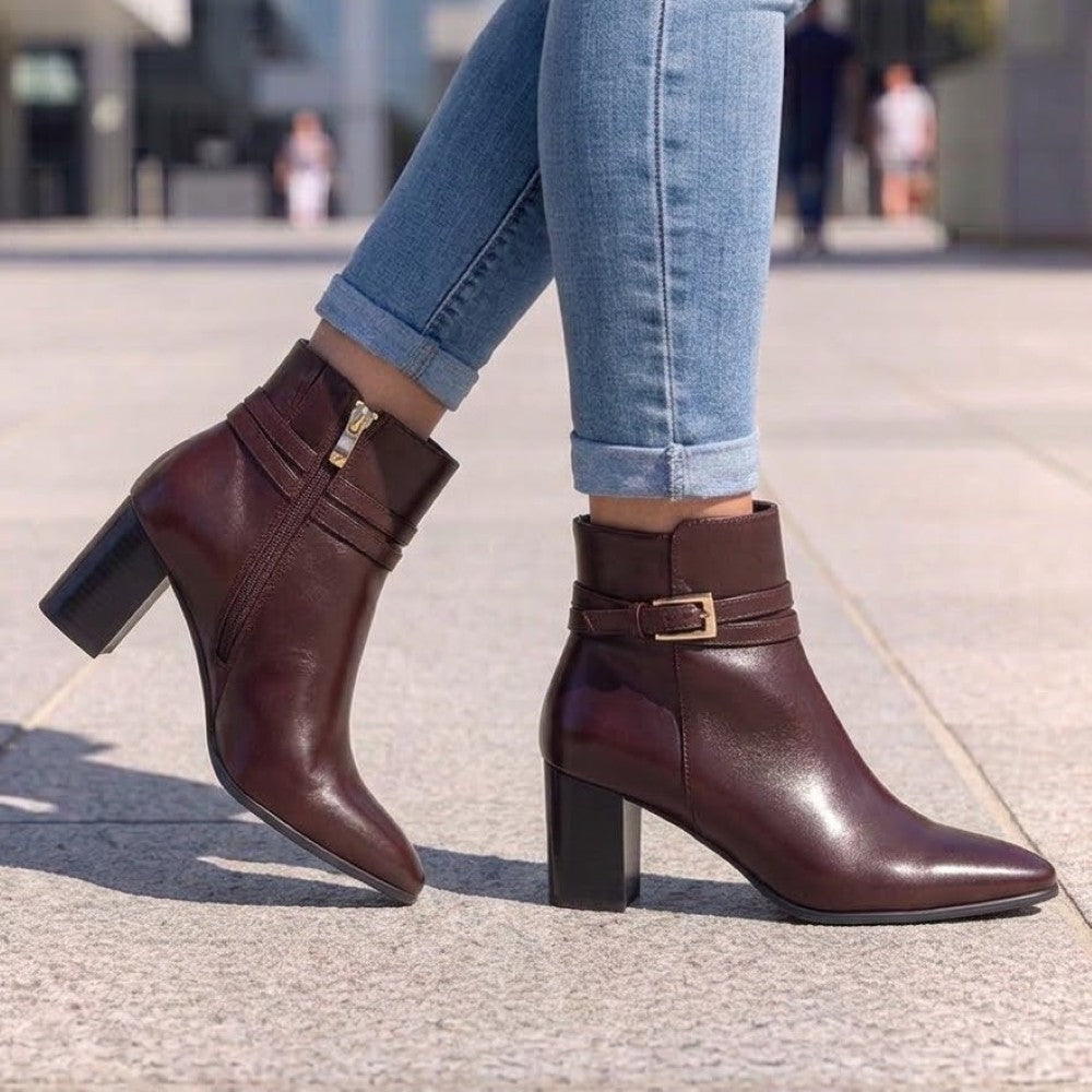 A pair of caprice dark brown leather ankle boots with block heels, worn with light blue cuffed jeans on a paved outdoor walkway. One boot is shown from the side with a zipper, and the other shows the front with a decorative strap and gold buckle.
