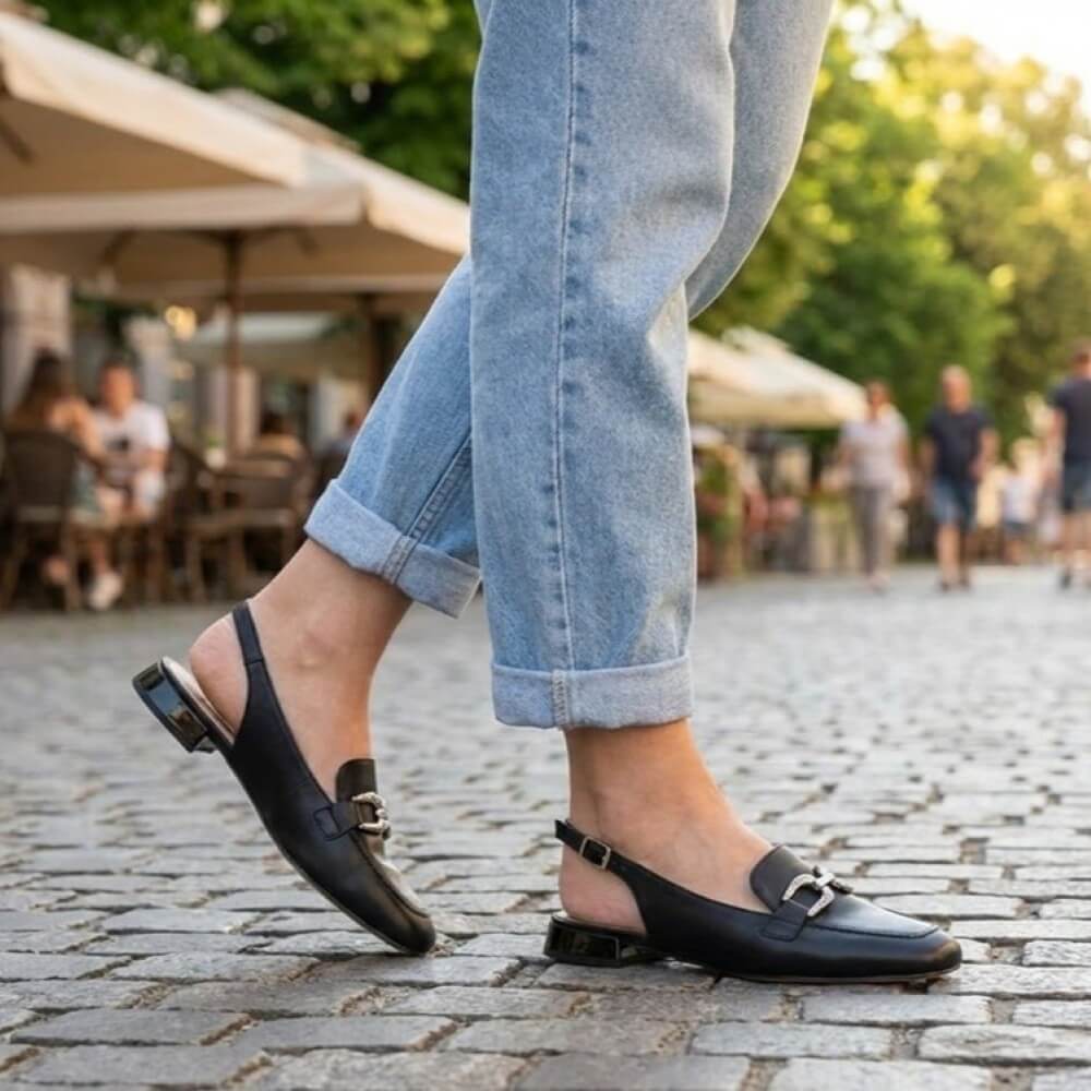 Person wearing Caprice 29400 black flat shoes and light blue jeans on a cobblestone street.
