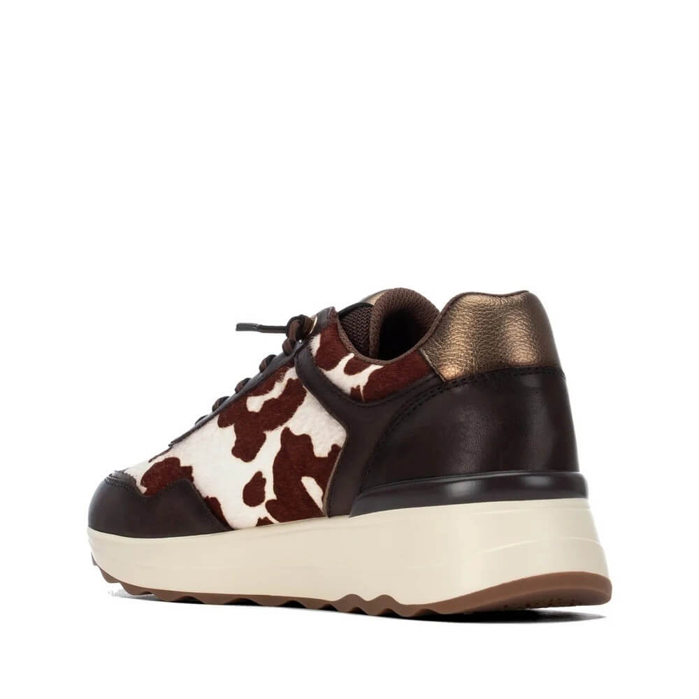 A low-top platform sneaker featuring dark brown leather, a brown and white cow-print pattern, a metallic bronze heel tab, and a thick cream-colored platform sole.