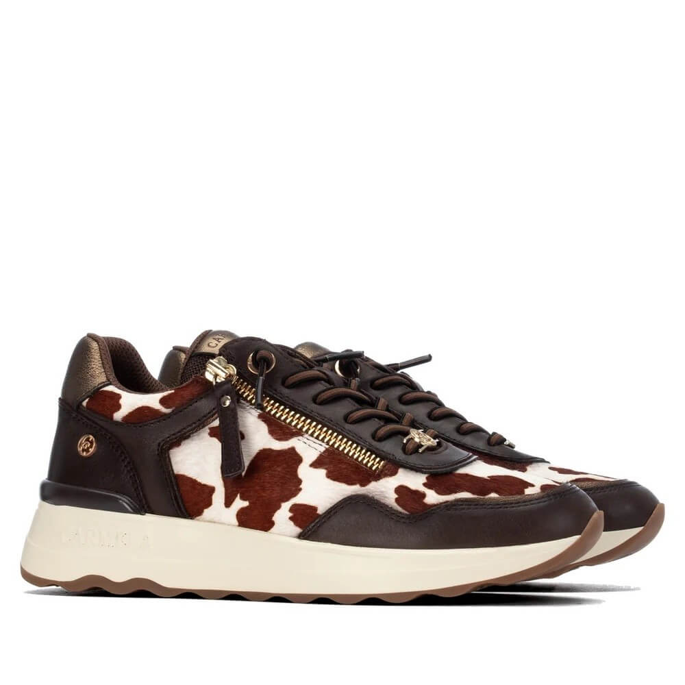 A pair of Carmela low-top sneakers featuring a brown and white cow-print pattern, dark brown leather overlays, a gold-tone side zipper, and a thick cream-colored sole. The brand name 'Carmela' is embossed on the side of the sole.