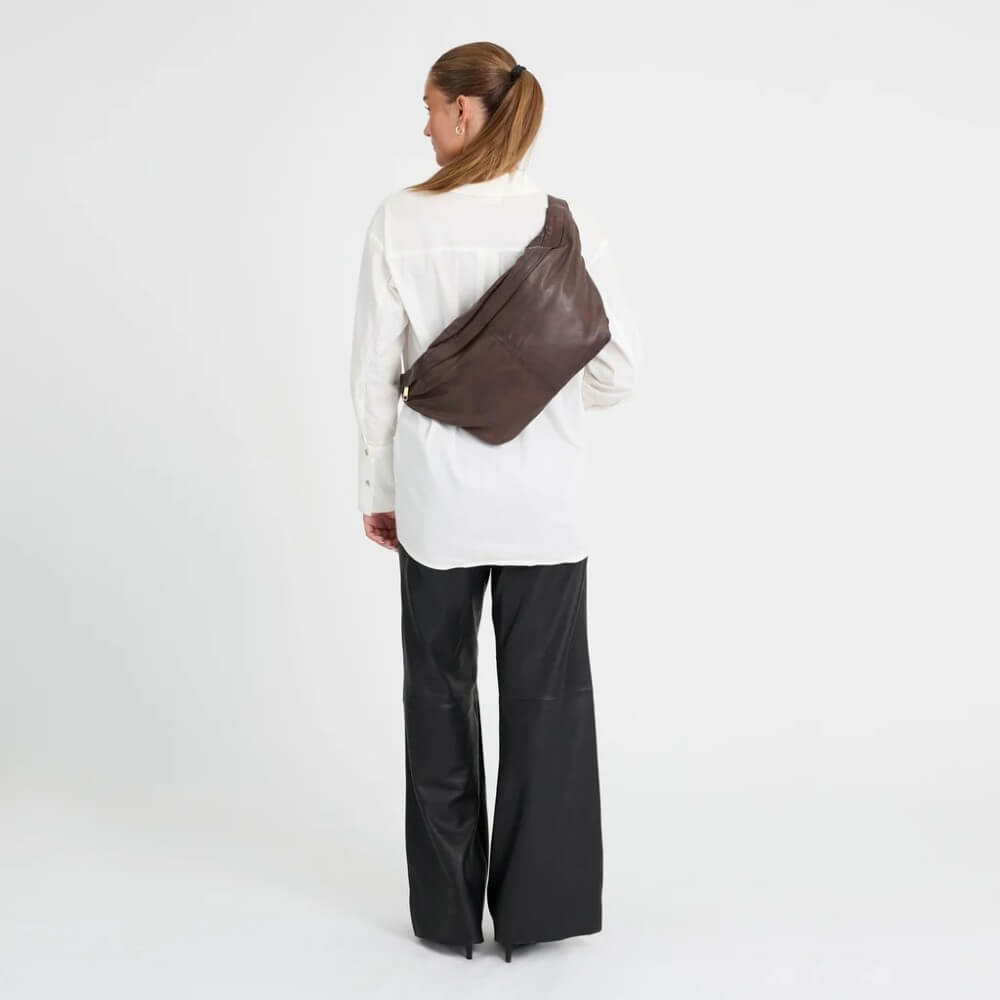 A woman viewed from behind wearing a white long-sleeve shirt, black wide-leg pants, and a dark brown leather sling bag across her back, set against a plain white studio background.
