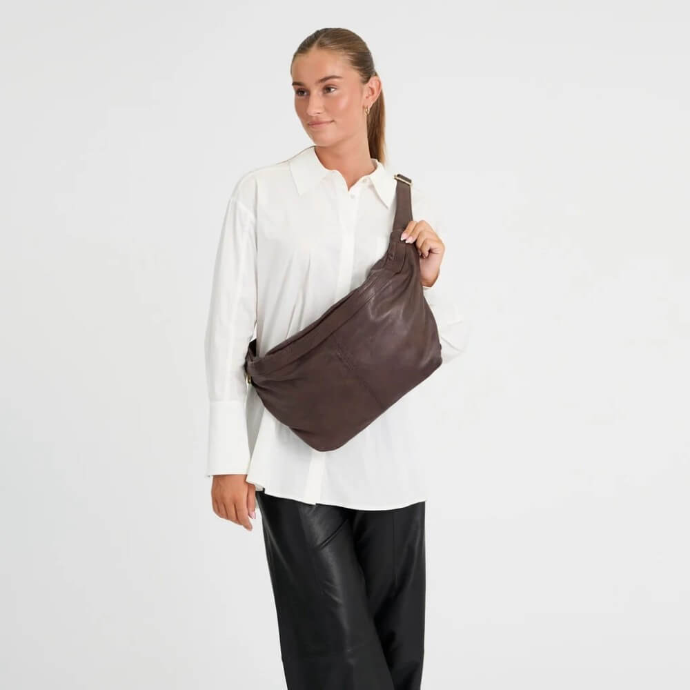 A woman wearing a white button-up shirt and black pants models a Depeche dark brown leather slouchy shoulder bag against a light grey studio background.