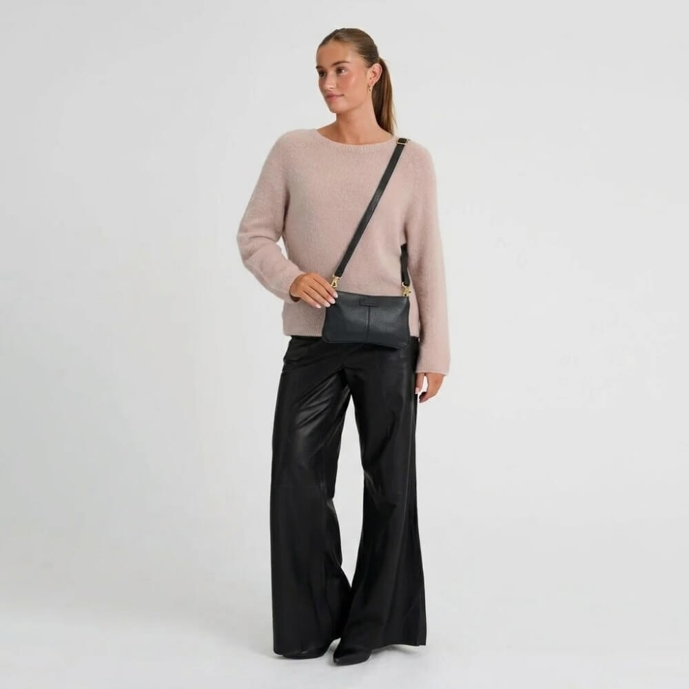 A woman modelling a light pink knit sweater, black wide-leg pants, and a Depeche black crossbody bag with gold hardware against a plain white studio background.