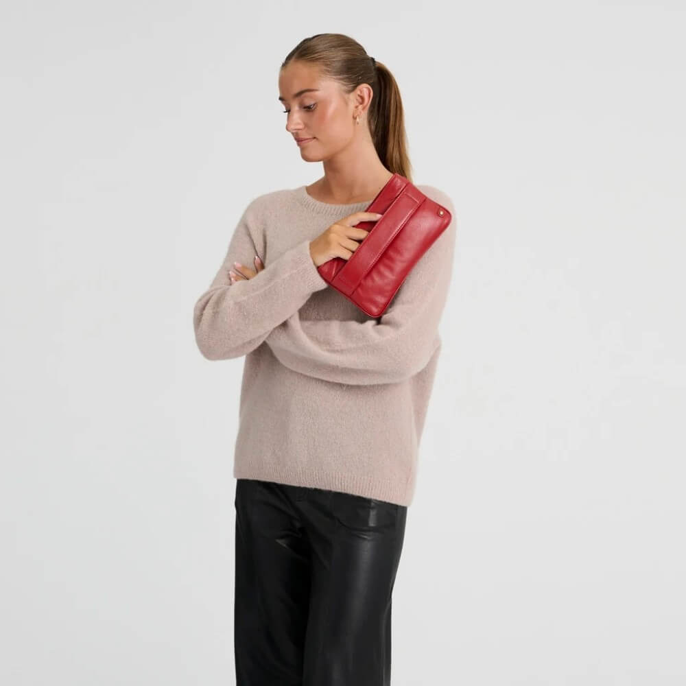 A woman wearing a beige knit sweater and black pants holds a Depeche red leather clutch bag against her chest, photographed against a light grey background.