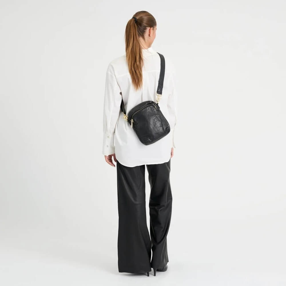 A woman wearing a white long-sleeve shirt and black pants is shown from the back, wearing a Depeche black leather crossbody bag with gold-tone hardware and a black strap against a plain white studio background.