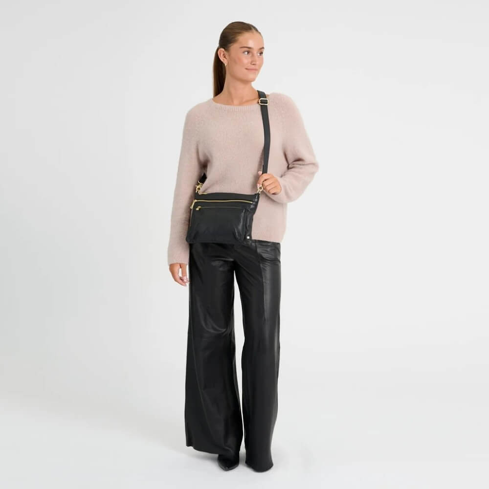 A woman wearing a light pink knit sweater and black pants is shown from the side, wearing a Depeche black leather crossbody bag with gold zippers and hardware against a plain white studio background.
