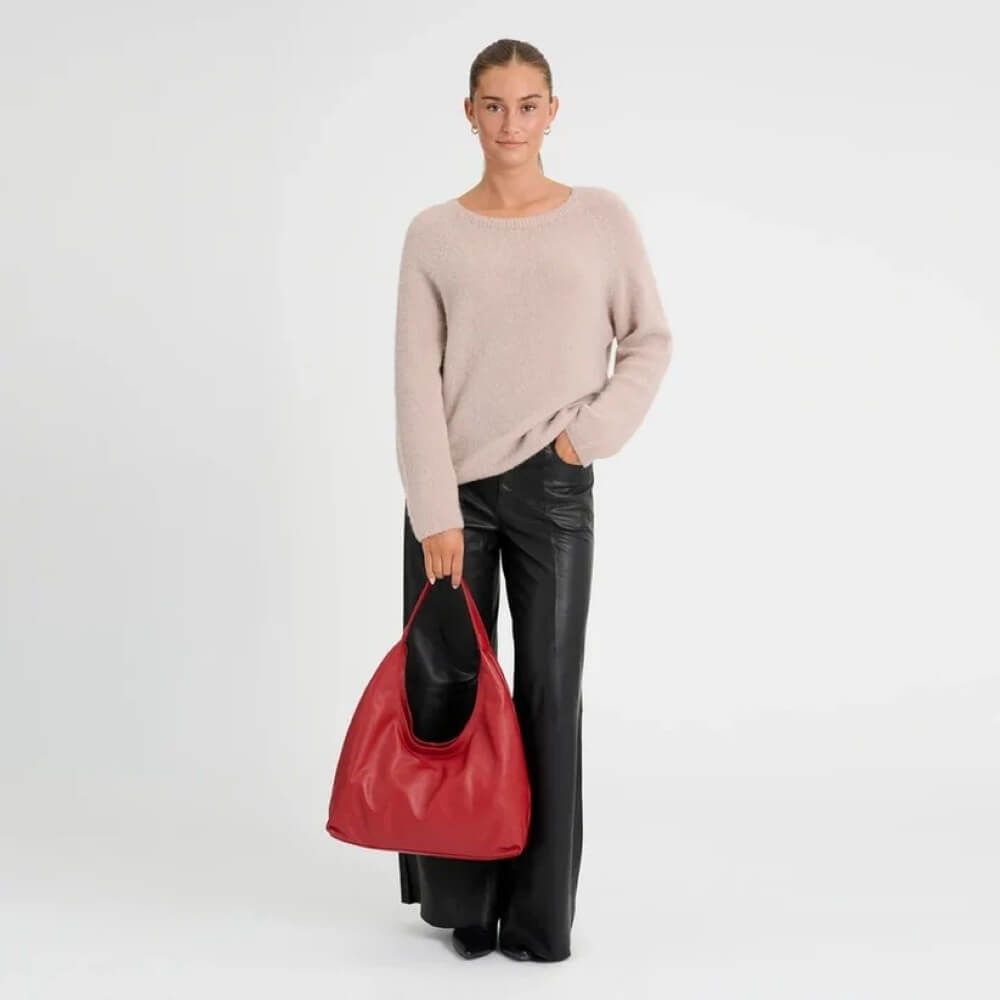 A woman in a light pink sweater and black pants carries a large Depeche red leather shoulder bag against a plain white studio background.