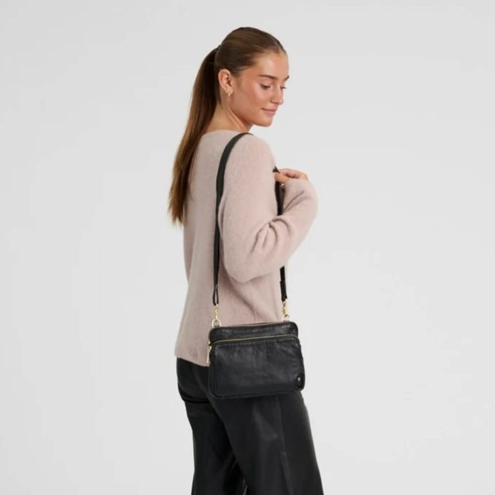 A woman wearing a light pink knit sweater and black pants is shown from the back and side, carrying a Depeche black leather crossbody bag with gold-tone hardware against a plain light grey studio background.