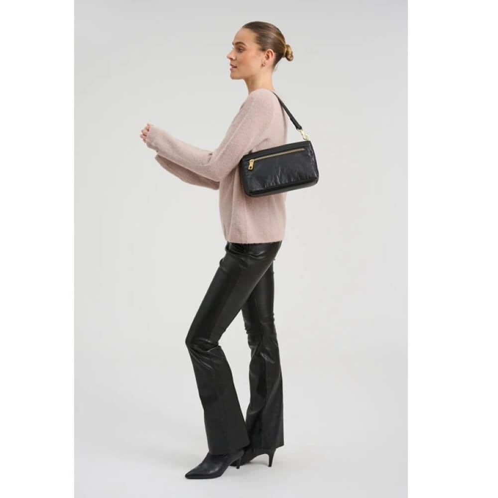 Woman wearing a light pink sweater and black leather pants, holding a black handbag on a white background